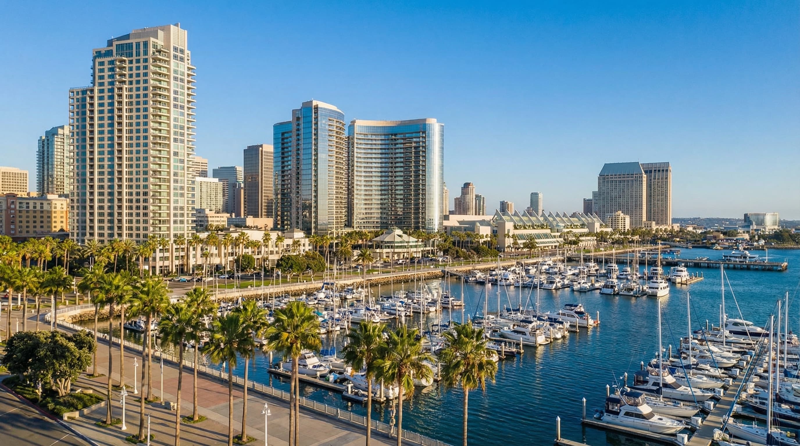 Downtown San Diego California waterfront skyline with modern high-rise buildings, palm trees lining Embarcadero, and sailboats in marina on a bright sunny day