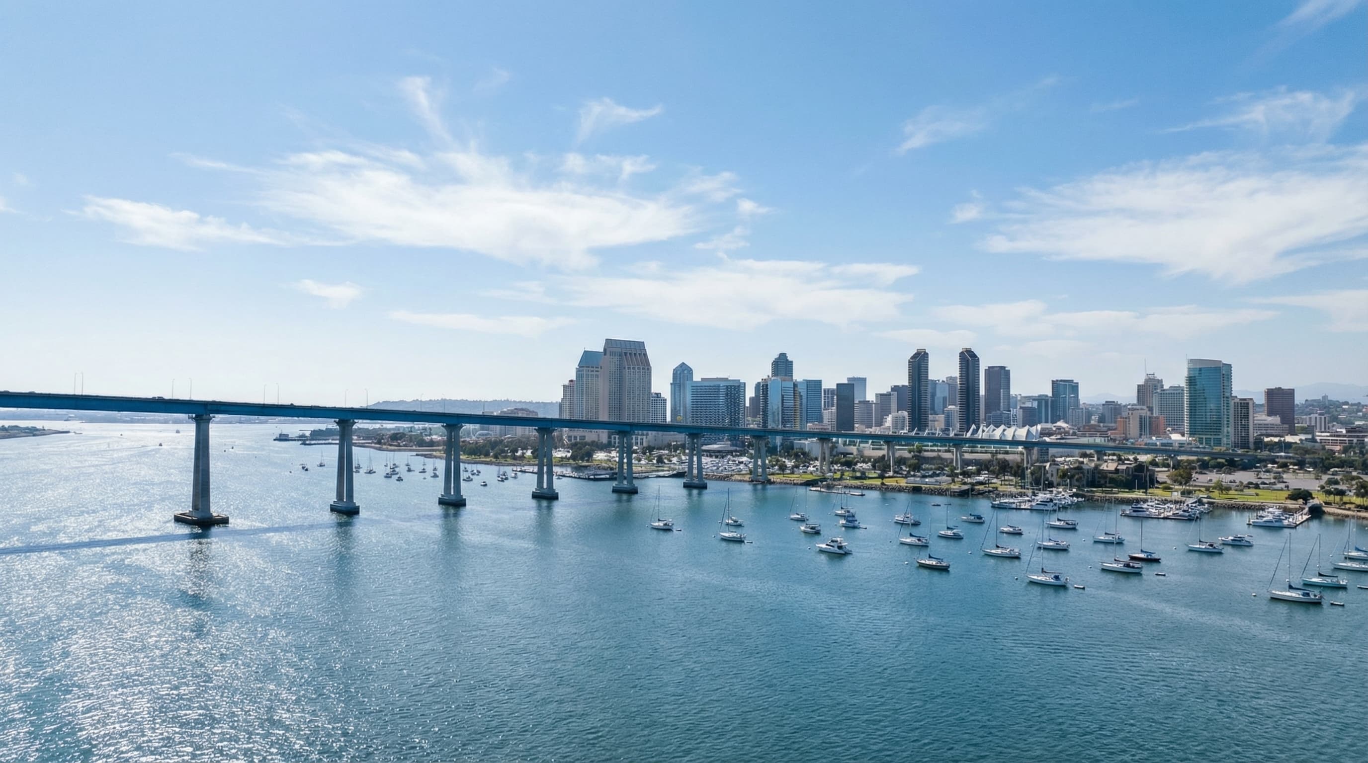 Aerial view of San Diego skyline from Coronado Bay with iconic Coronado Bridge spanning across bay, sailboats in harbor, and clear blue California sky
